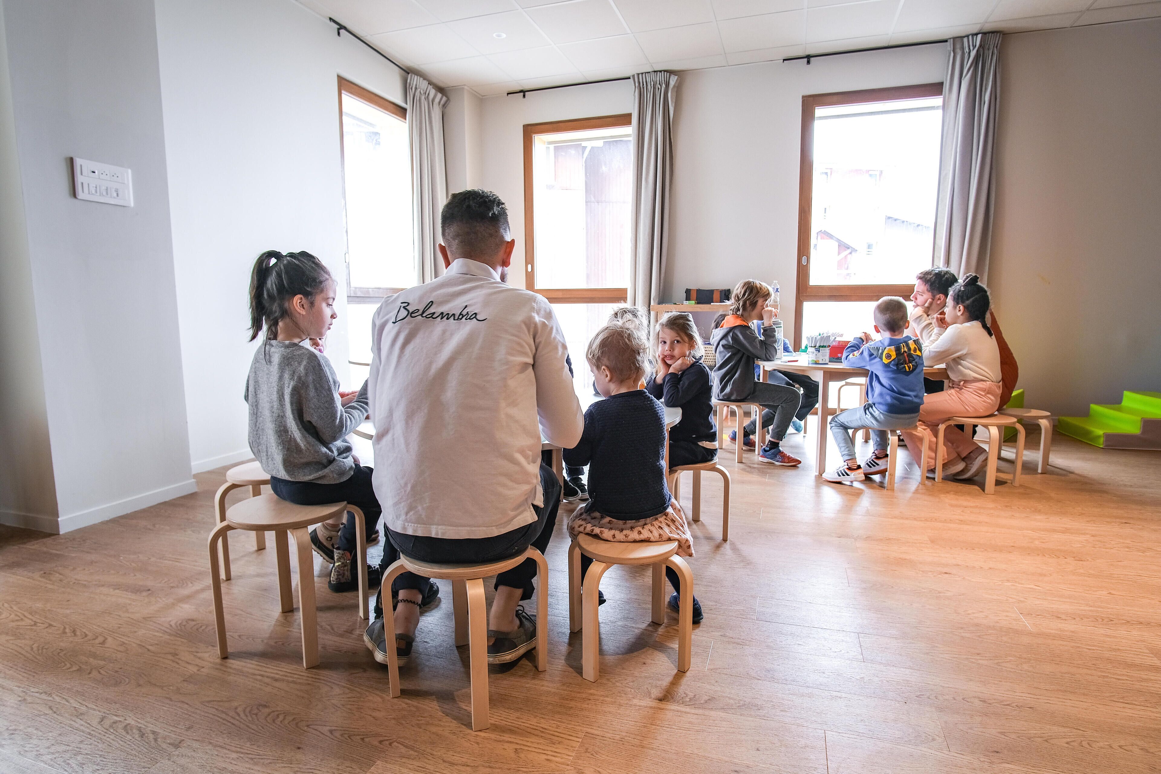Een groep volwassenen en kinderen zitten aan tafels in een lichte ruimte met grote ramen en houten vloeren.