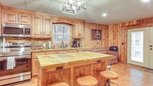 A kitchen with wooden cabinets, a large island with stools, stainless steel appliances, and a hanging light fixture. - Wooded Blue Ridge Cabin: 2 Decks, Fire Pit! (Blue Ridge)