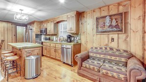 A kitchen with wooden cabinets, a stainless steel dishwasher, a bar stool area, and a patterned sofa. - Wooded Blue Ridge Cabin: 2 Decks, Fire Pit! (Blue Ridge)