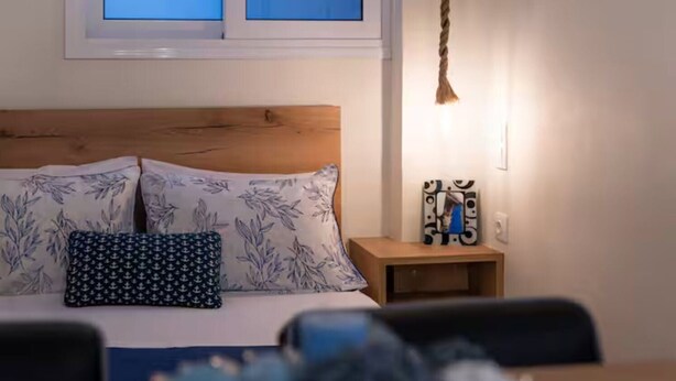 A bedroom with a wooden headboard, a bed with floral and anchor-patterned pillows, a bedside table with a decorative item, and a hanging light fixture. - Flare Studio  (Corfu)