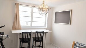 Ein Zimmer mit einem weißen Schreibtisch, zwei schwarzen Stühlen, einem Fahrrad, einer Chandelüster, einem fenster mit Vorhängen und einem leeren Bilderrahmen an der Wand.