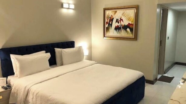 Una cama bien hecha con ropa de cama blanca y azul, una mesita de noche con una lámpara y un cuadro abstracto enmarcado en la pared.