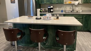 Una cocina con armarios verdes, un refrigerador de acero inoxidable, una estación de café y tres taburetes de bar.