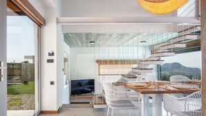 Un intérieur moderne comprenant un coin repas, des chaises en osier blanches, une table en bois et une grande fenêtre donnant sur l’extérieur.