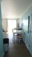 Un comedor con una mesa, sillas y una computadora. Hay una ventana con cortinas, un cesto de basura y un cuadro en la pared.