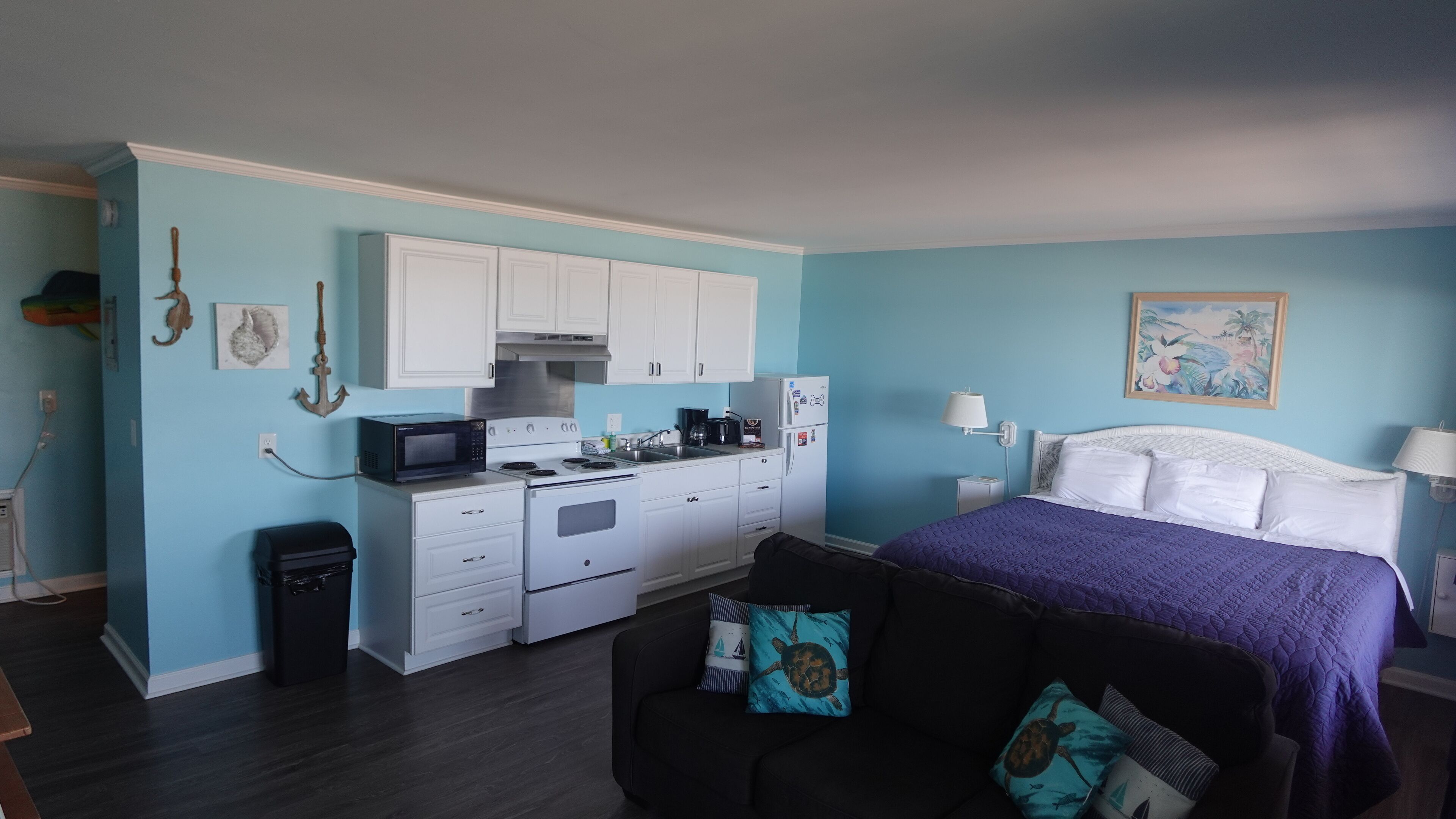 A compact room with a bed, a sofa, a kitchenette, and a small trash bin.
