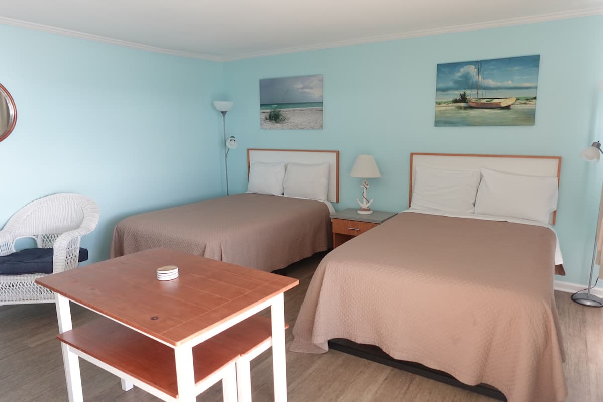 A hotel room with two beds, a wooden table, a wicker chair, and artwork on the walls.