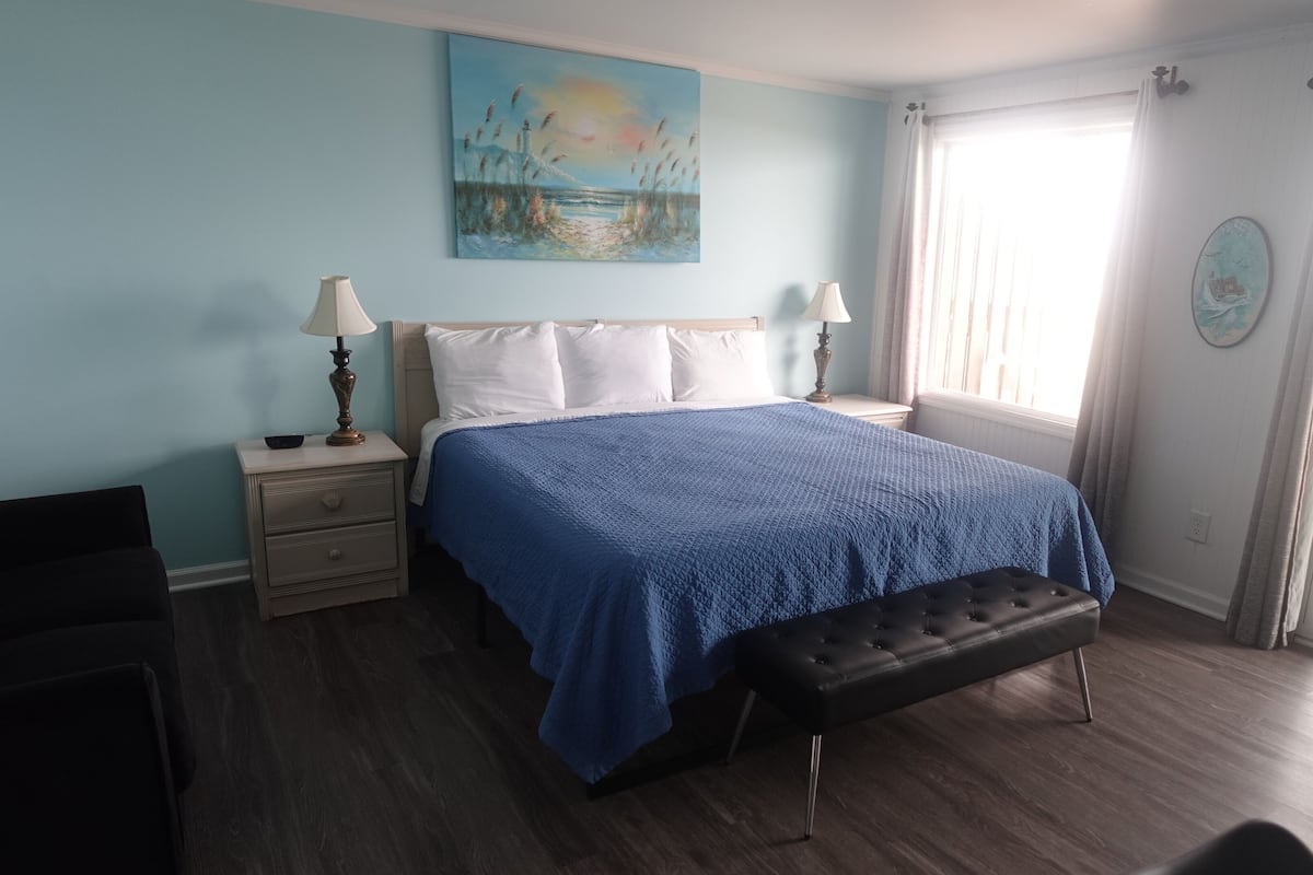 A bedroom with a bed, bedside tables, a bench, a painting, and a window with curtains.