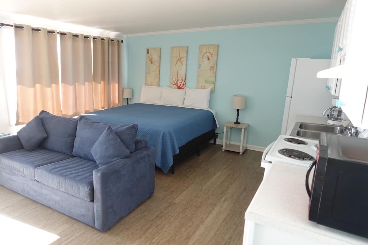 A bedroom with a bed, a sofa, a nightstand, a lamp, and a kitchenette.