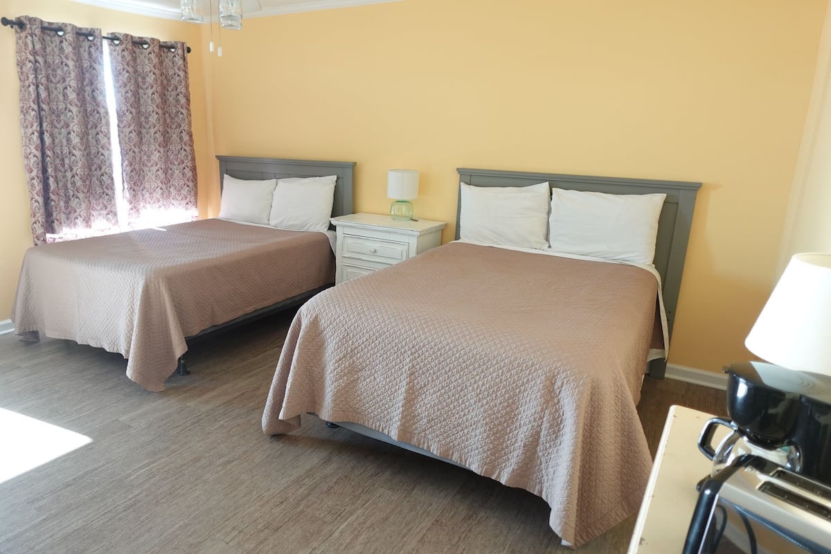 A hotel room with two beds, a nightstand, a lamp, and a window with curtains.