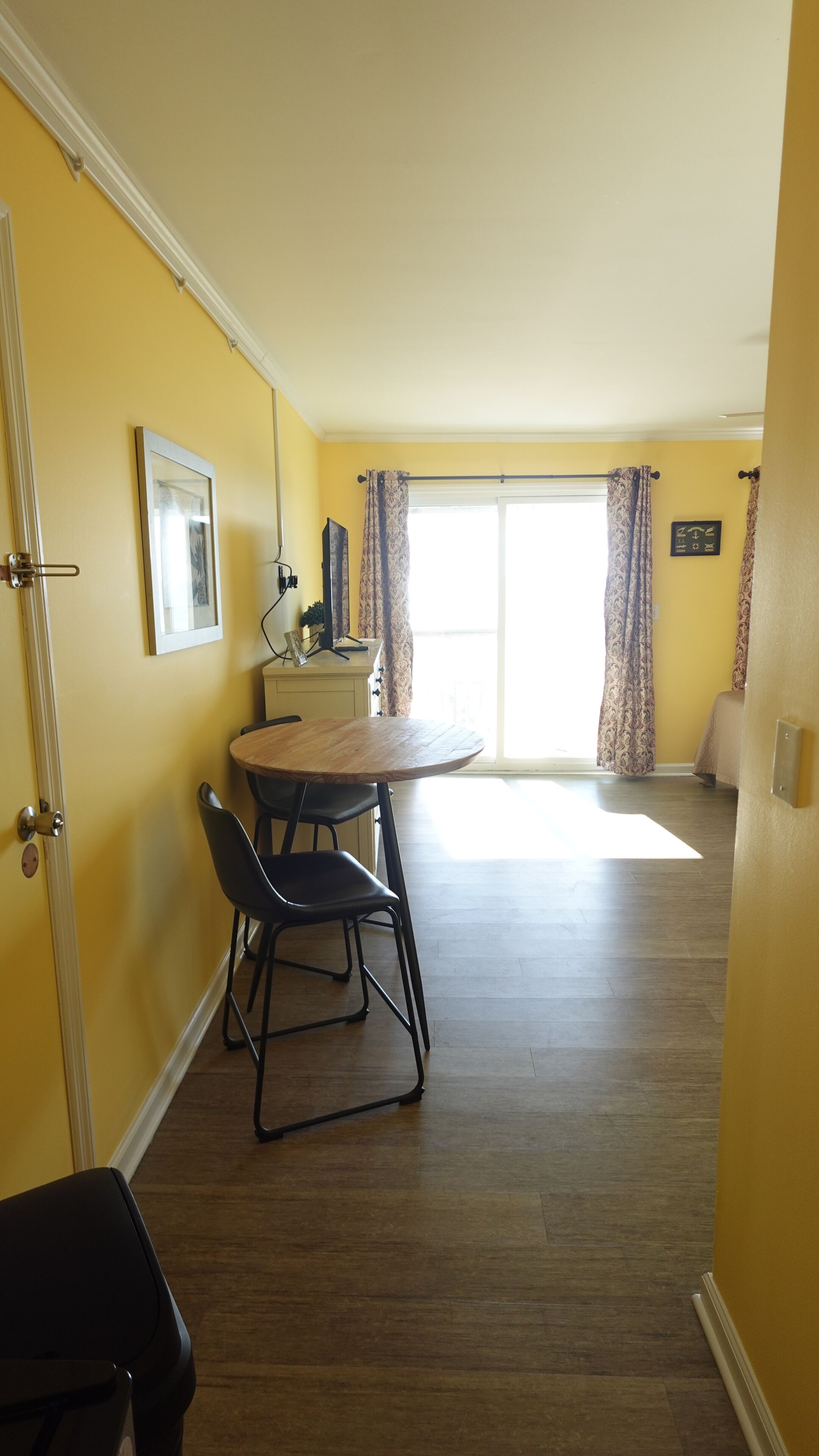 A room with a dining table and chairs, a bed, a window with curtains, and a door.