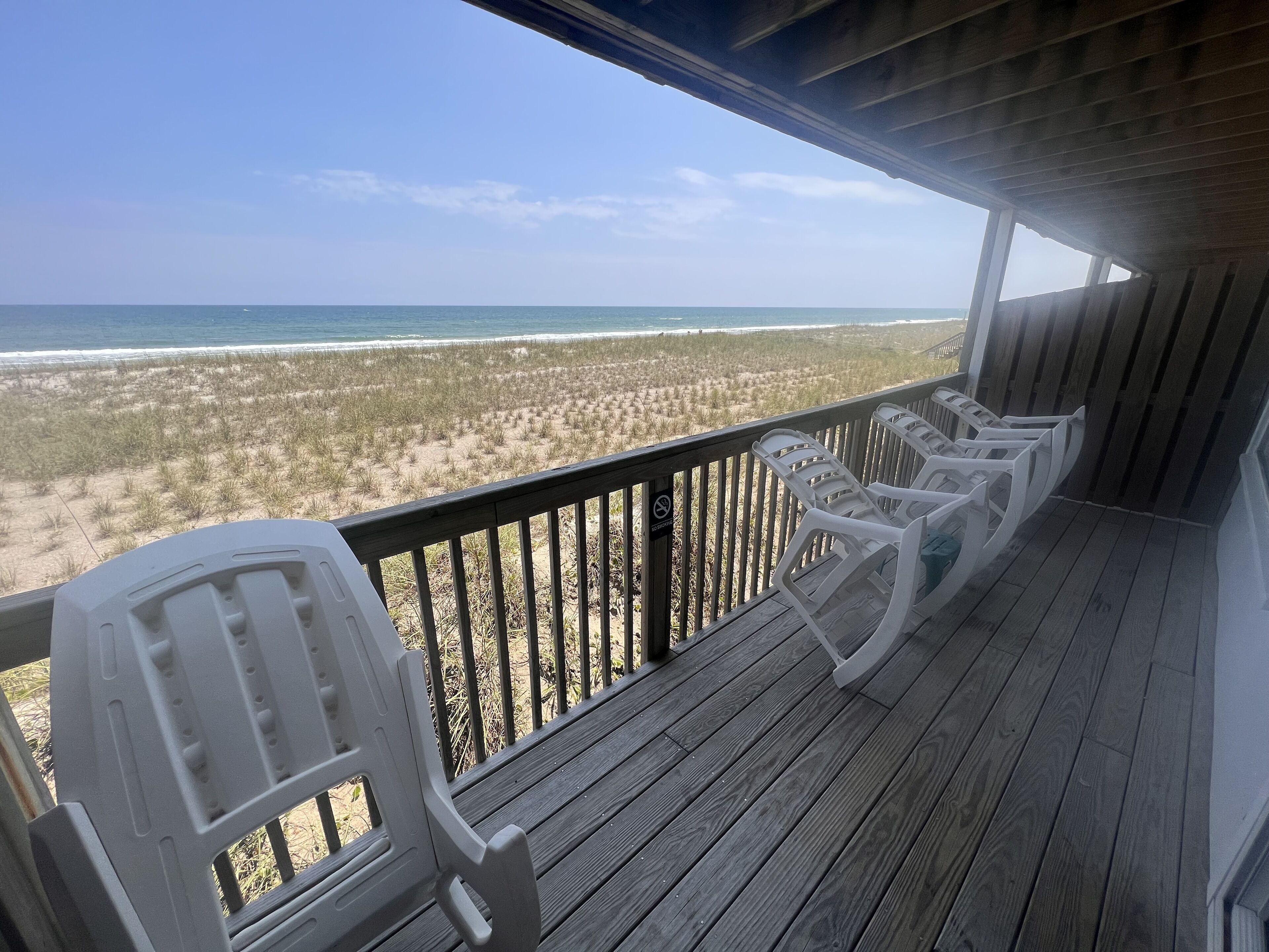 Een houten terras met witte kunststof stoelen met uitzicht op een strand.