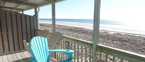 A wooden deck with two turquoise Adirondack chairs facing a beach.