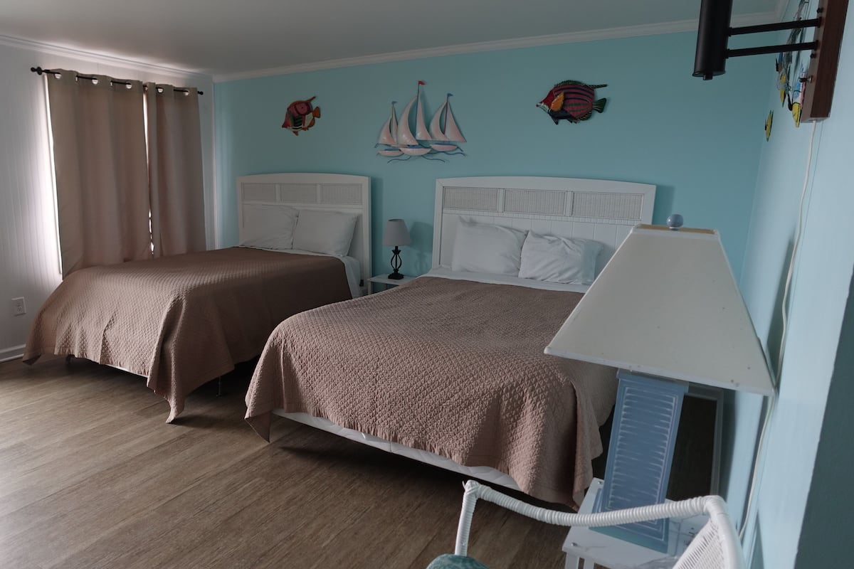 A double room with two beds, a window with curtains, a wall with decorative fish and sailboats, and a white chair.