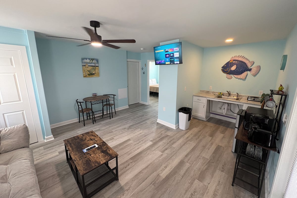 A living area with a ceiling fan, a TV mounted on the wall, a dining table with chairs, a coffee table, and a kitchen area with a sink and countertop.