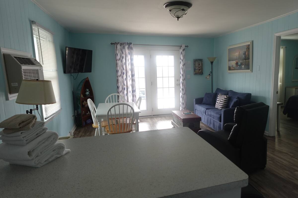 A room with a dining table, chairs, a sofa, and a TV. There is a window with blinds and a door leading to another room.