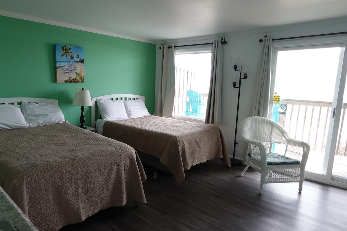 A hotel room with two beds, a green wall, a painting, a chair, and a balcony with a view.