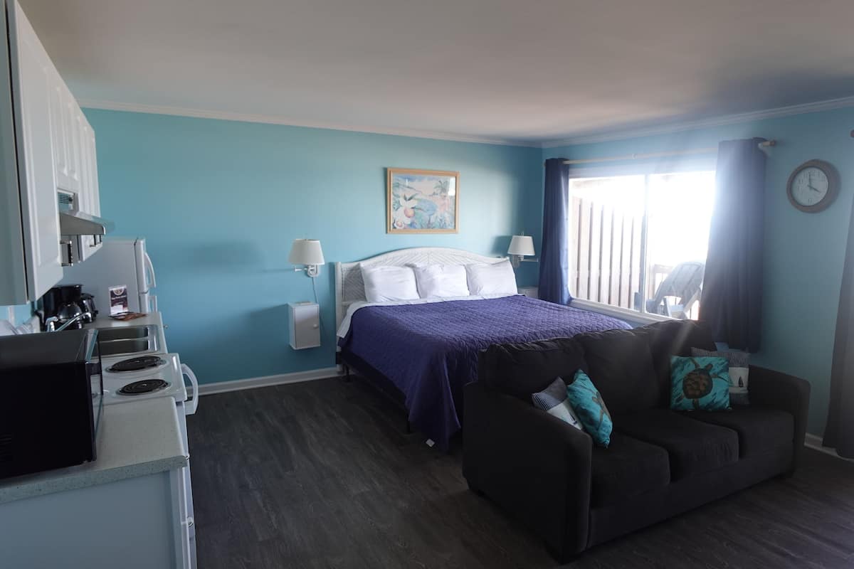 A hotel room with a bed, a sofa, a kitchenette, and a window with curtains.