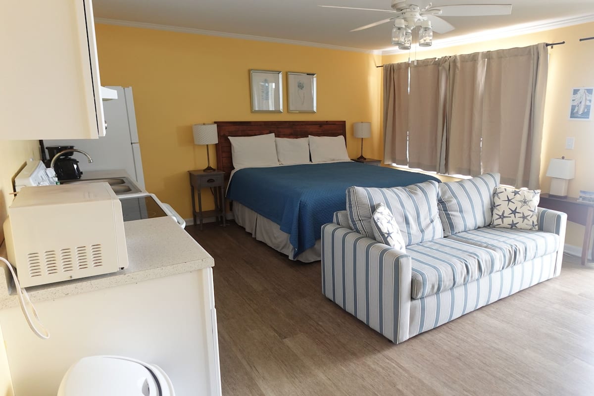 A hotel room with a bed, a sofa, a ceiling fan, a window with curtains, and a kitchenette area.