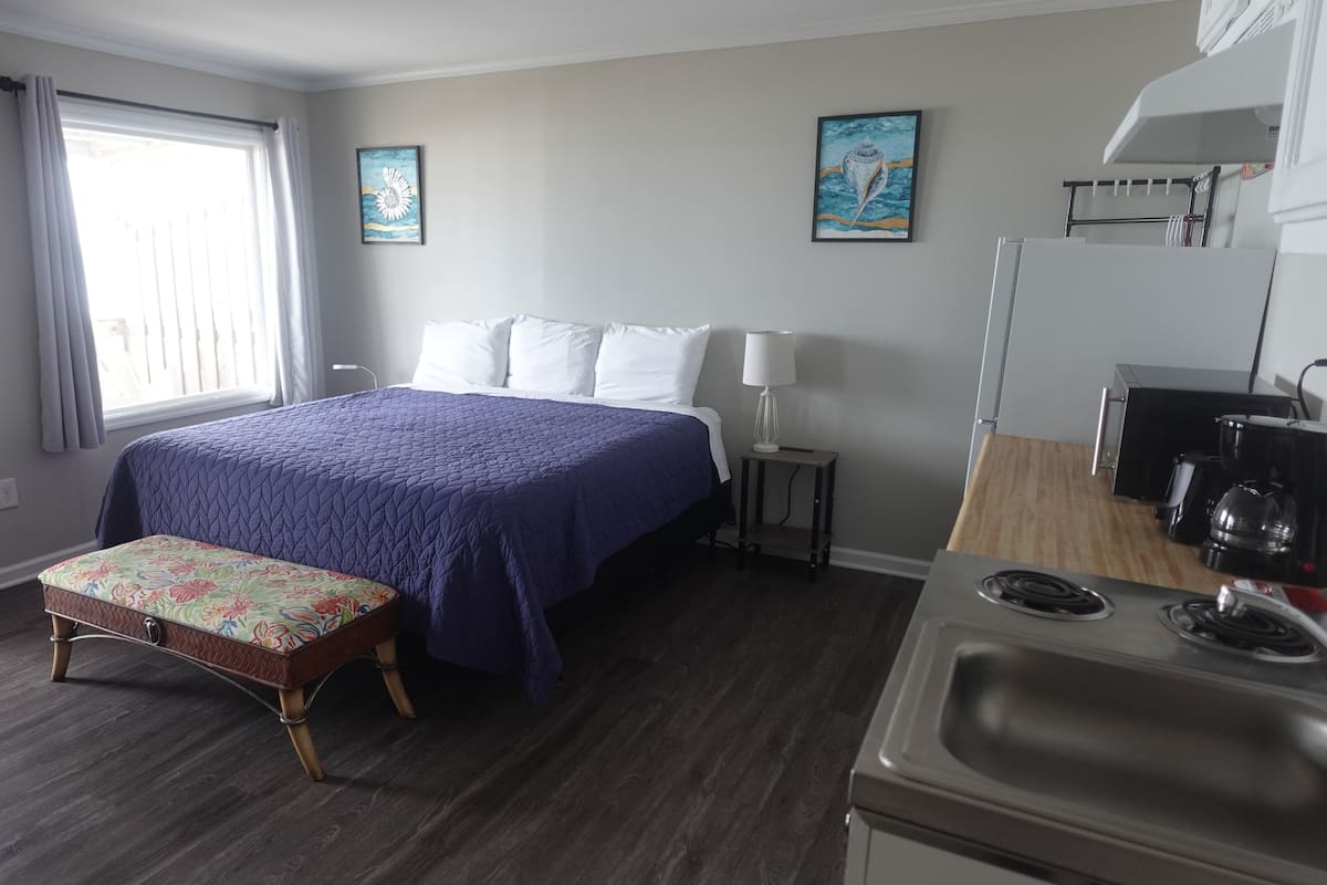 A bedroom with a bed, a bench, a nightstand, a lamp, and a kitchenette.