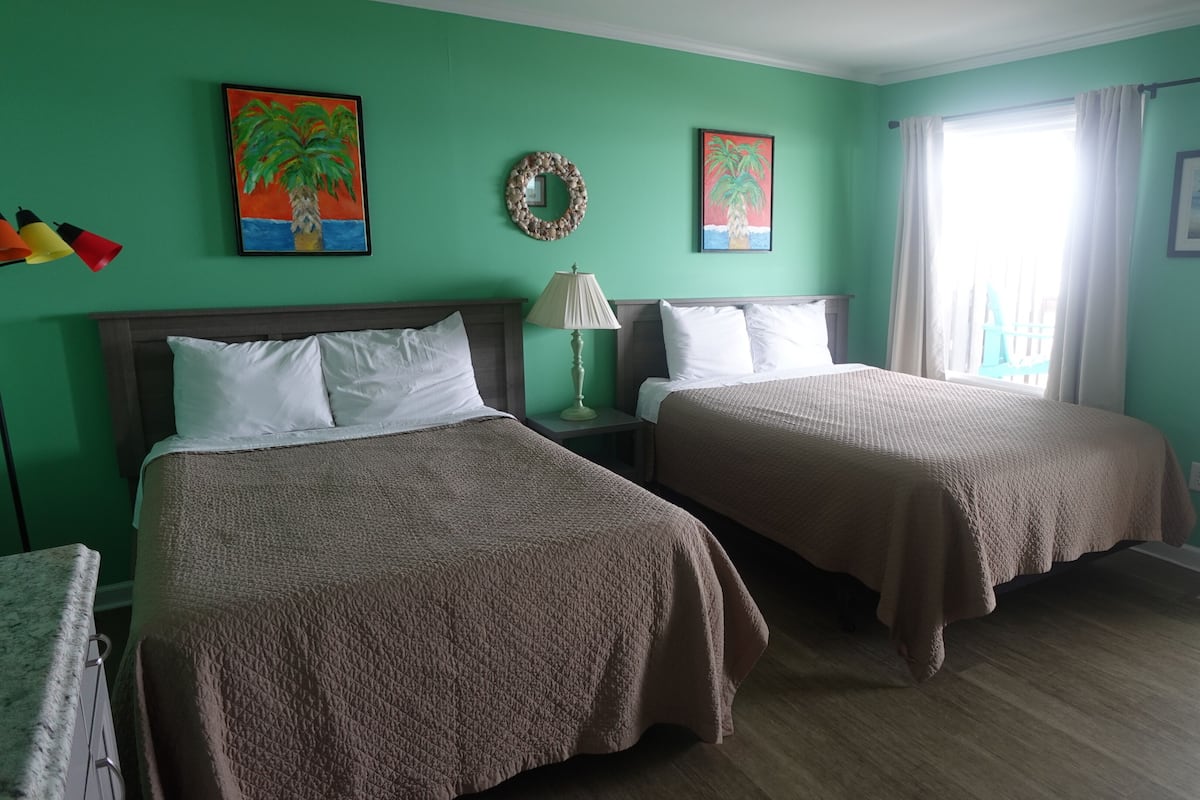 A room with two beds, a nightstand with a lamp, a wall painting, and a window with curtains.