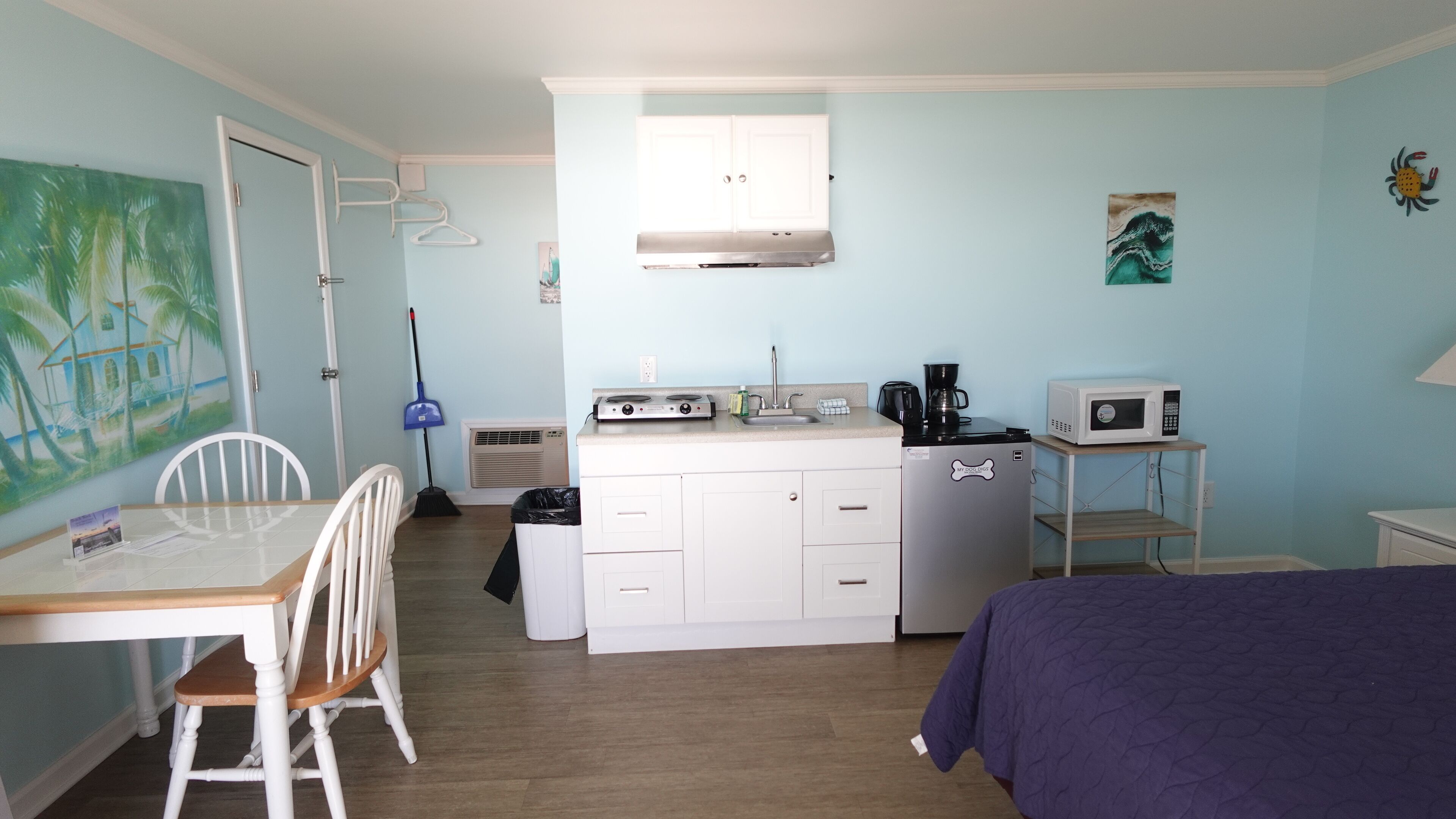 A compact kitchenette with a microwave, coffee maker, and a small refrigerator. There is a dining table with chairs, a bed, and a painting on the wall.