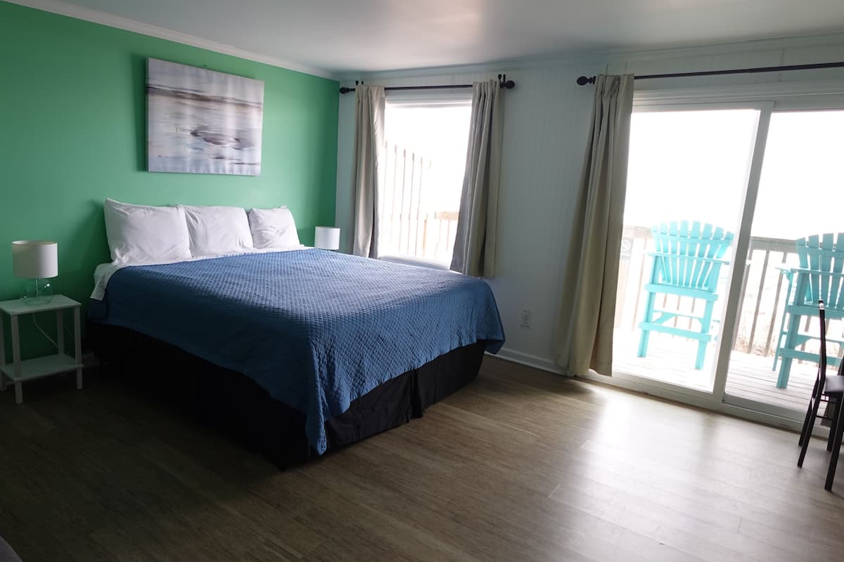 A bedroom with a bed, a nightstand, a lamp, a window with curtains, and a view of outdoor chairs.