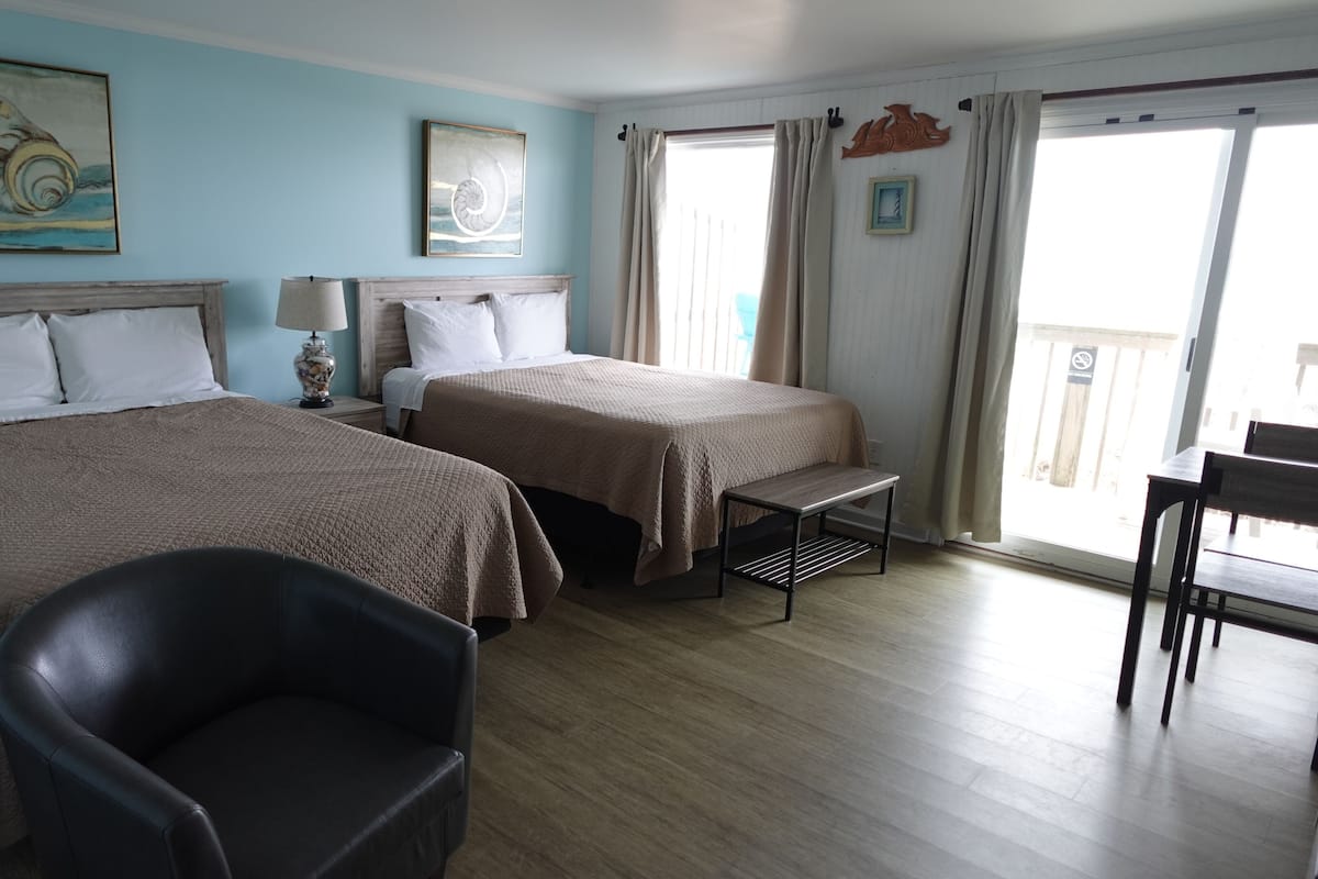 A hotel room with two beds, a chair, a small table, and a balcony with curtains.