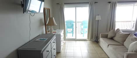 A room with a TV mounted on the wall, a sofa, a desk with drawers, a lamp, and a view of a balcony with outdoor furniture.