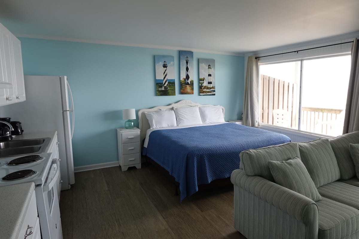 A bedroom with a bed, a nightstand, a dresser, a kitchenette, and a window with curtains.