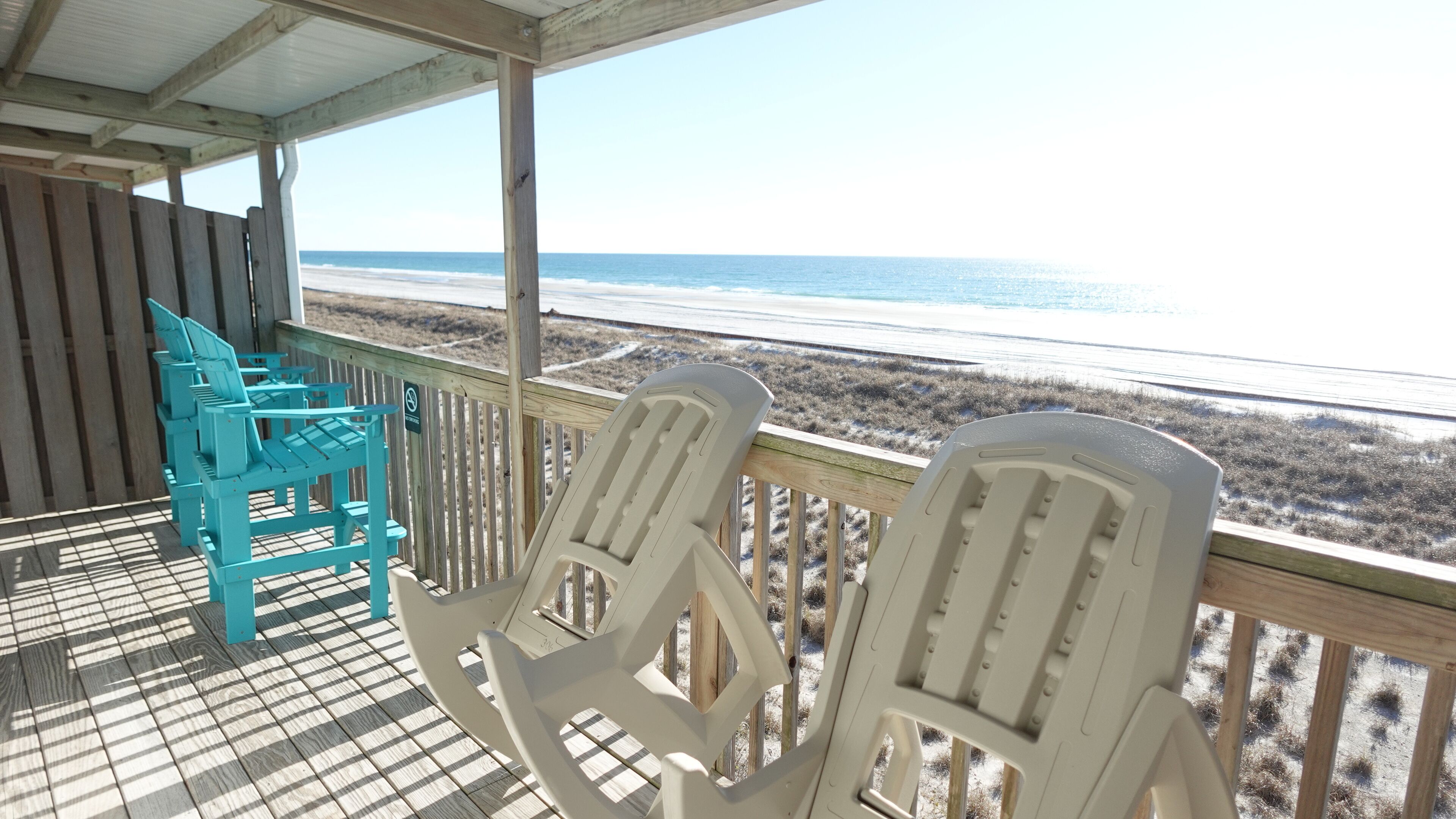 A covered deck with two white Adirondack chairs facing a beach.