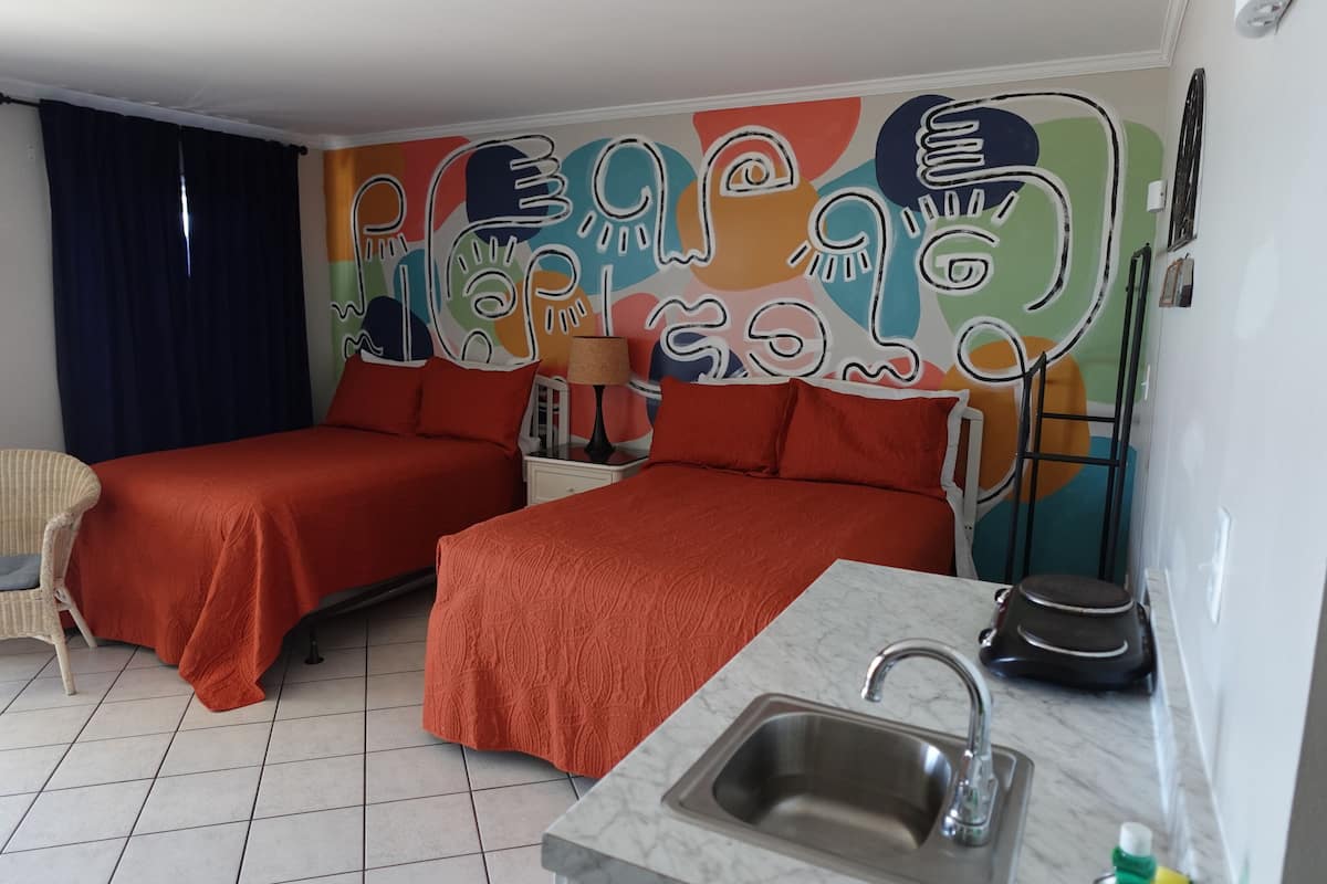 A room with two beds, a chair, a sink, and a colorful mural on the wall.