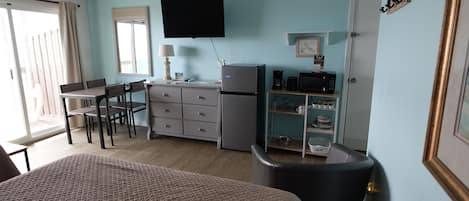 Een kleine kamer met een bed, een bureau met een stoel, een kleine koelkast, een magnetron en een plank met servies.