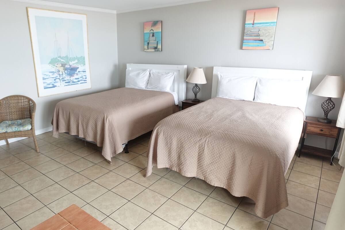 A hotel room with two beds, a wicker chair, and artwork on the walls.