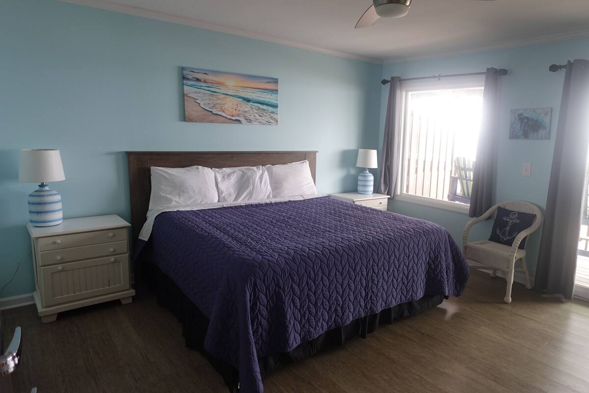 A bedroom with a large bed, two bedside tables with lamps, a chair, and a view of the outdoors.