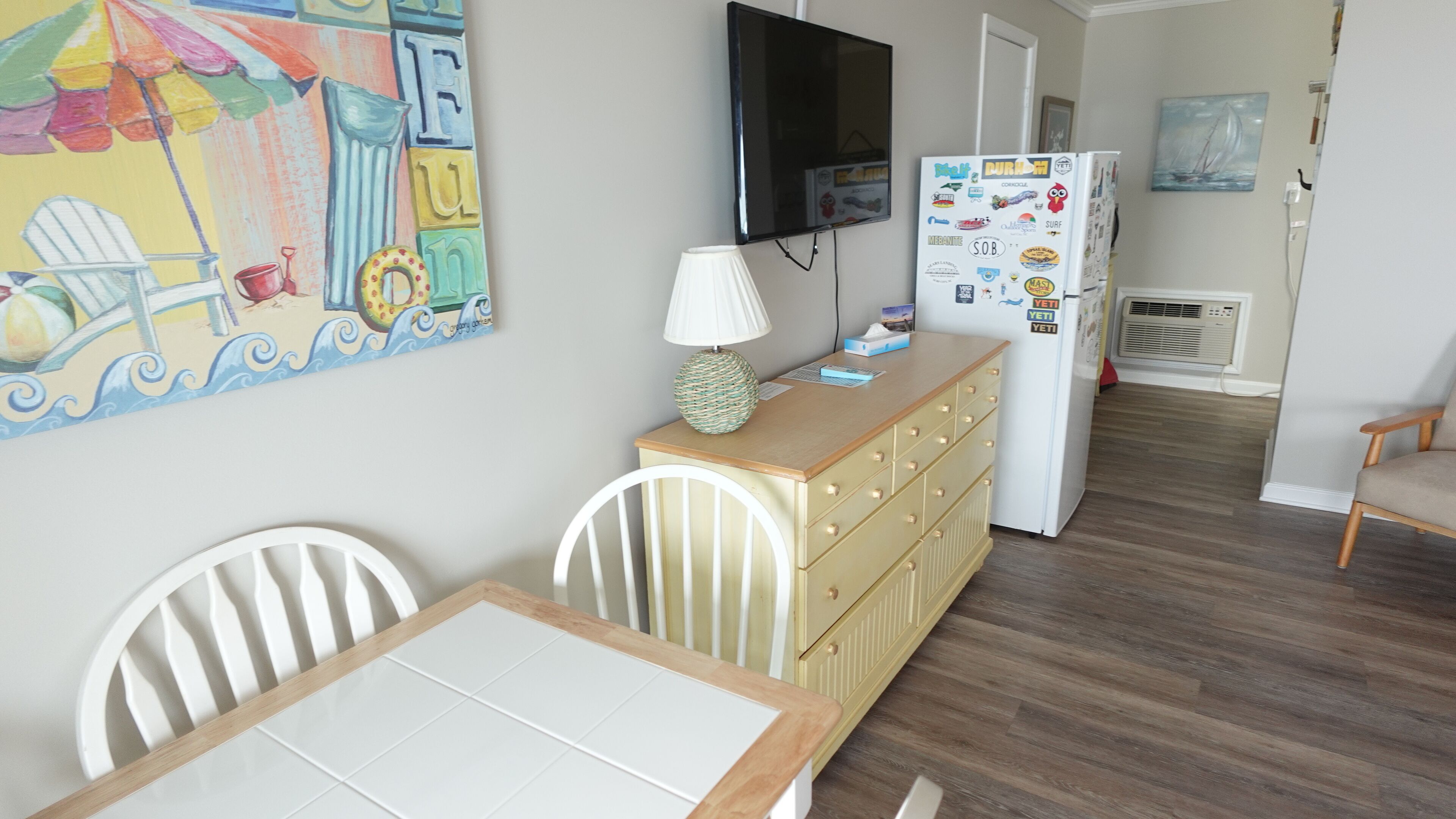 A dining area with a table and chairs, a lamp, a TV mounted on the wall, a refrigerator with magnets, and a painting on the wall.
