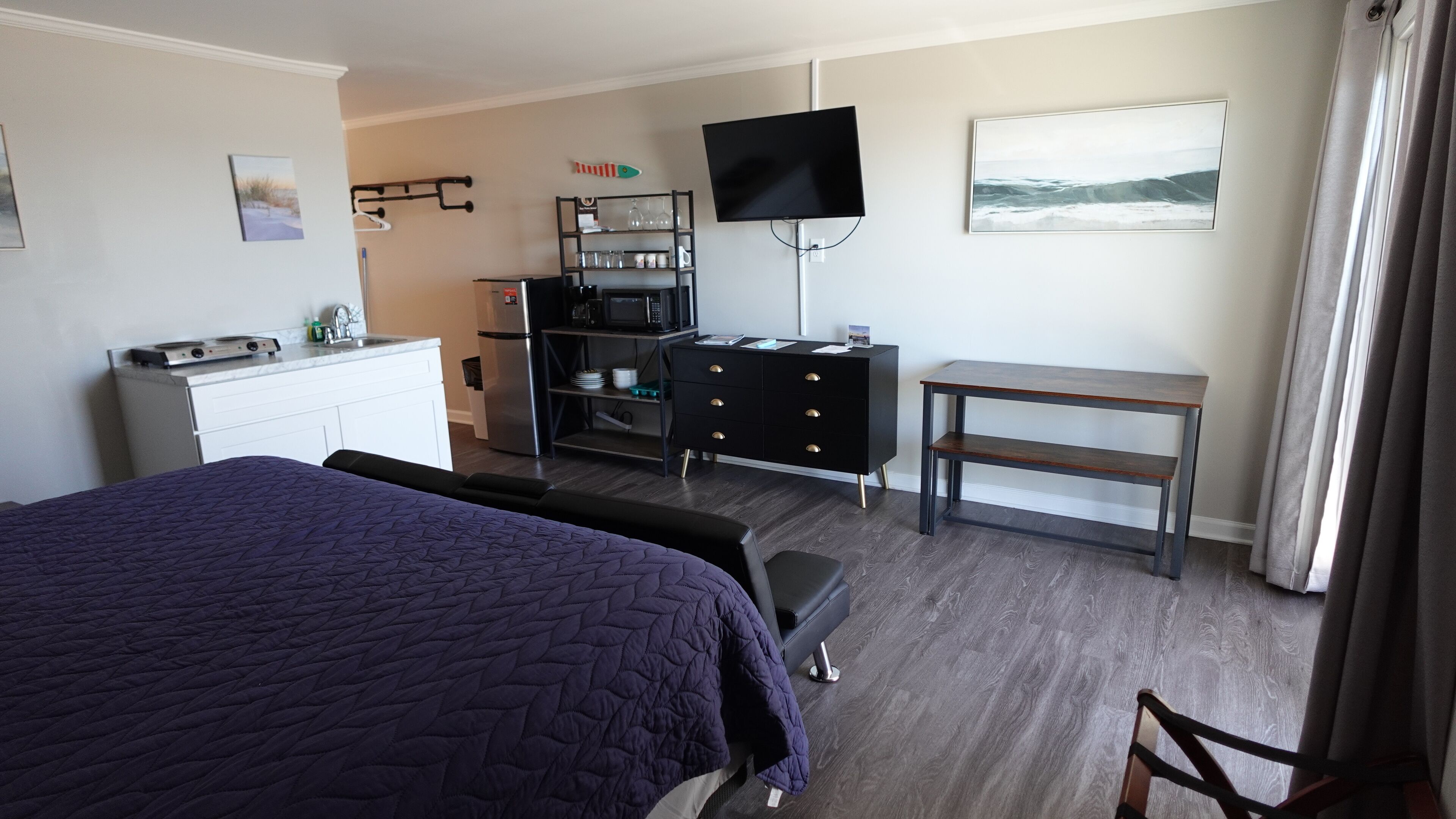 Habitación de hotel con cama, escritorio, televisión, kitchenette y ventana con cortinas.