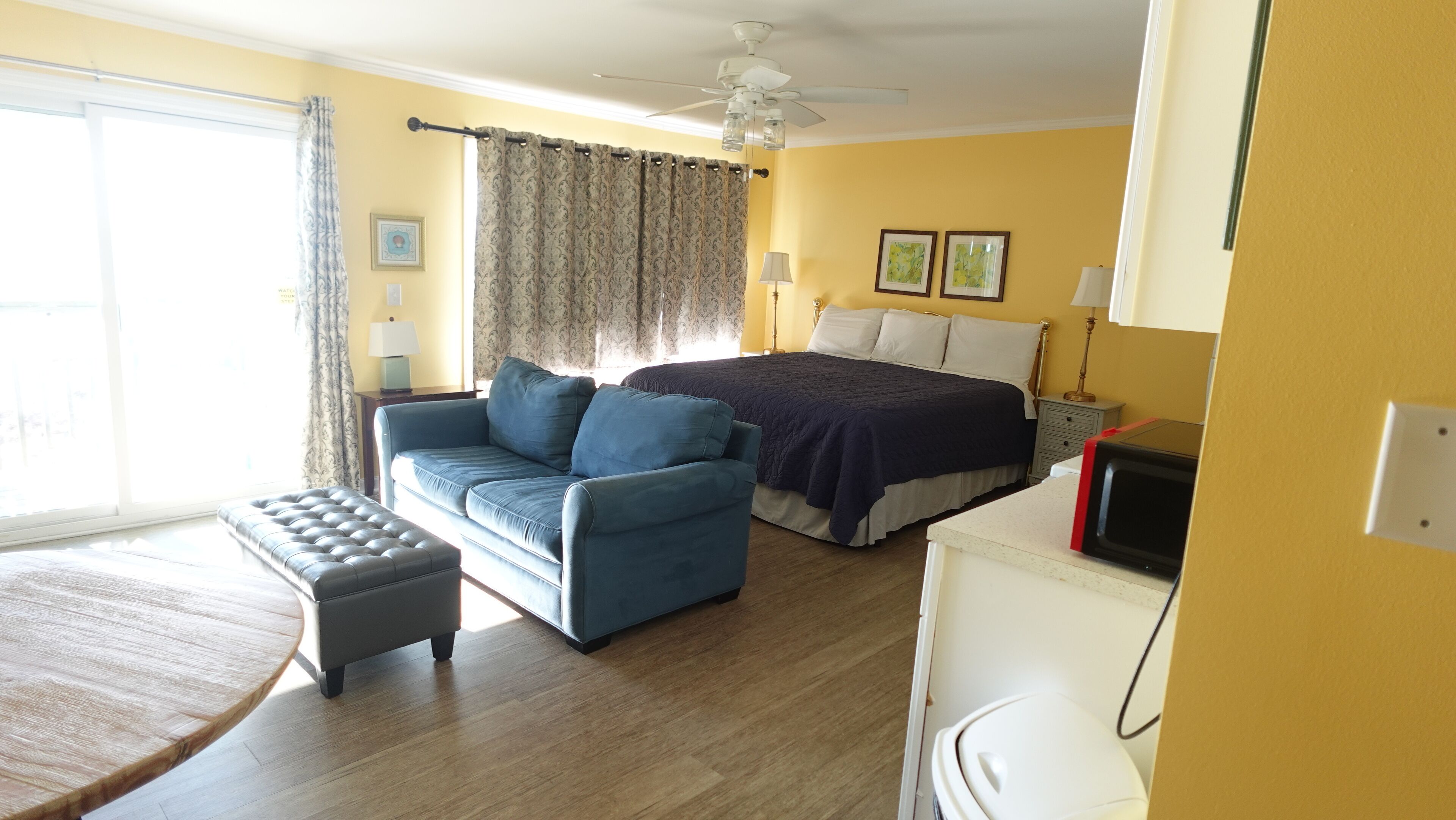 A hotel room with a bed, a sofa, a small table, a ceiling fan, and a television.