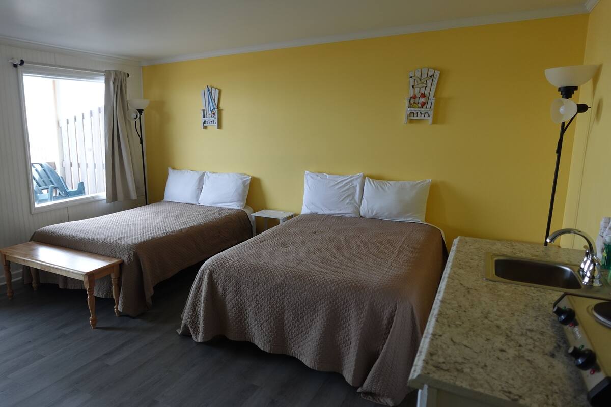A hotel room with two beds, a bench, a small table, a sink, and a window with curtains.