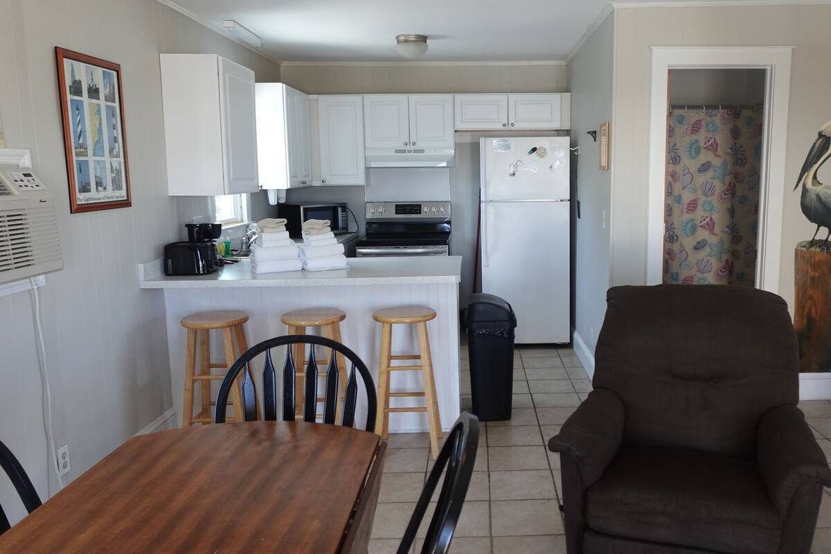 A compact kitchen with white cabinets, a refrigerator, a microwave, and a stove. There is a dining area with a wooden table and three stools. A trash bin is placed near the kitchen.