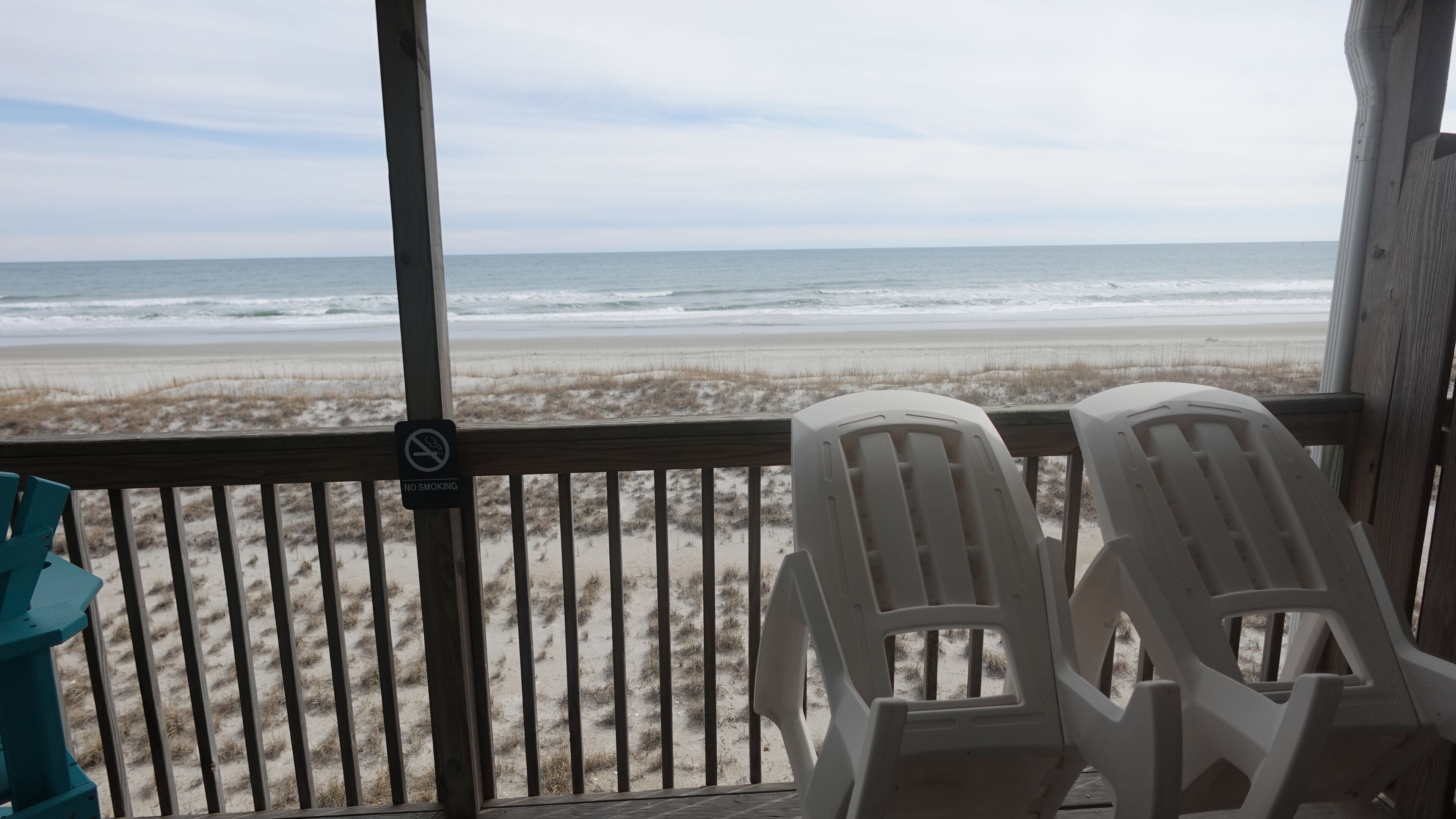 A beach view from a covered deck with two white plastic chairs and a 'No Smoking' sign.