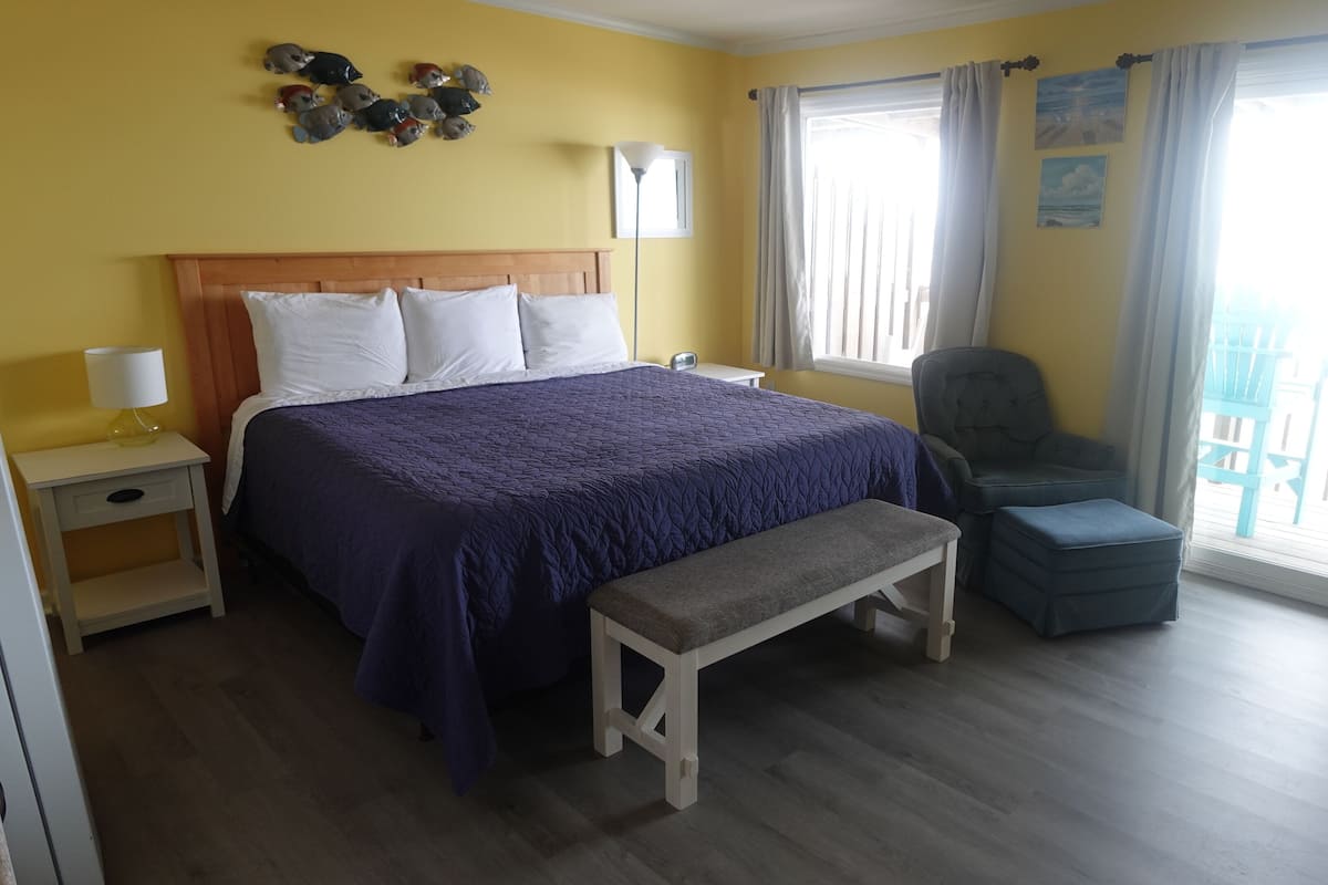 A bedroom with a bed, bedside table, bench, chair, and a balcony with a railing.