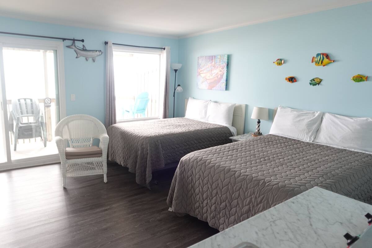 A room with two beds, a chair, a table, and a view of the outside.