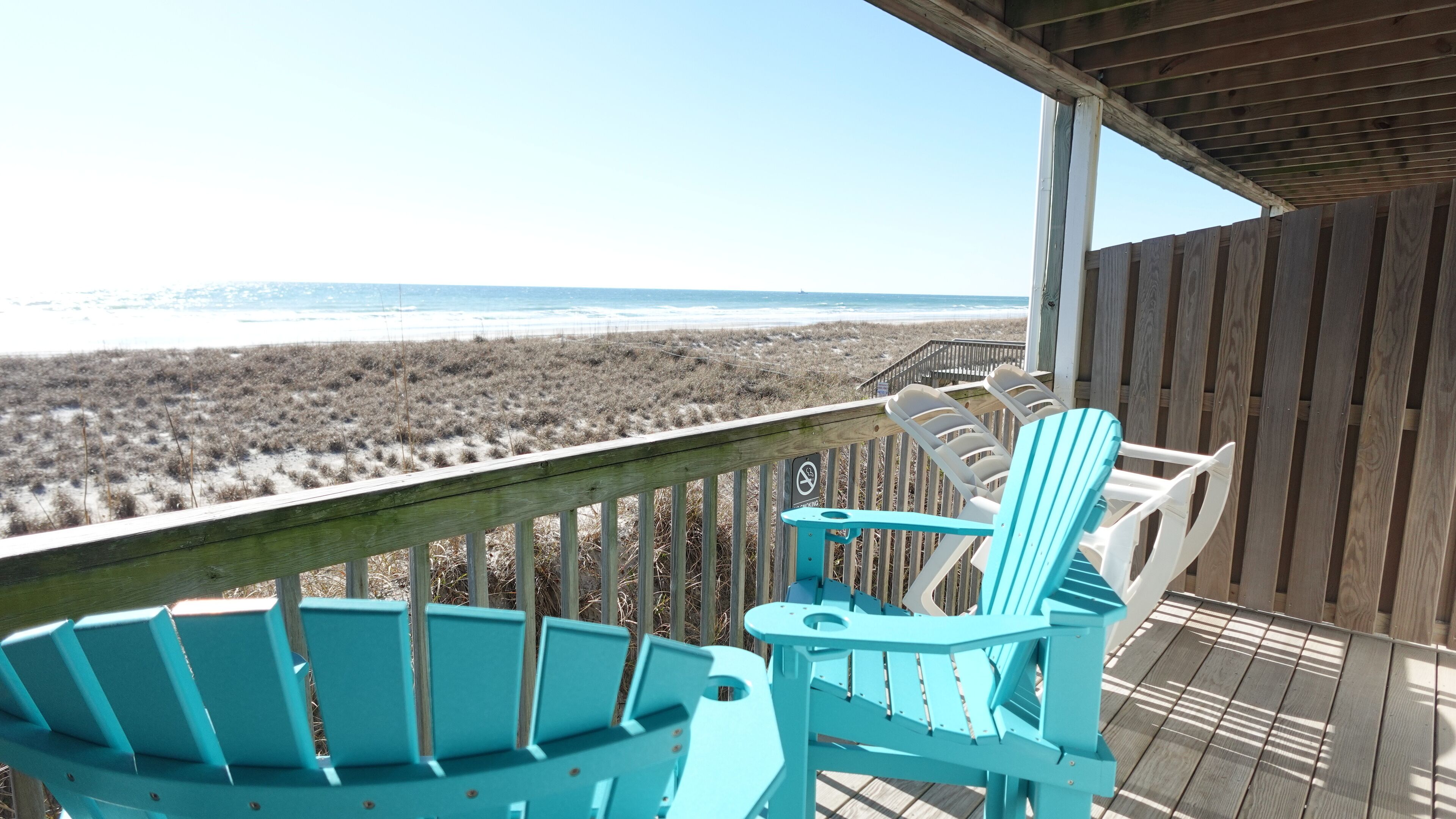 A wooden deck with two turquoise Adirondack chairs overlooking a beach.