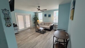 A living room with a beige sofa, a coffee table, a dining table with chairs, a ceiling fan, and a large window with blinds. - Sea Vista Motel (Holly Ridge)
