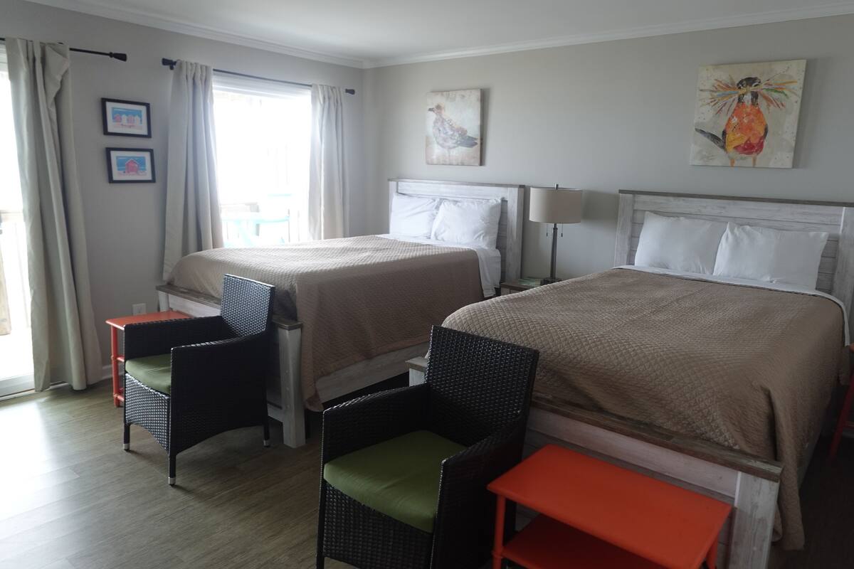 A hotel room with two beds, a window with curtains, a chair, a small table, and artwork on the walls.
