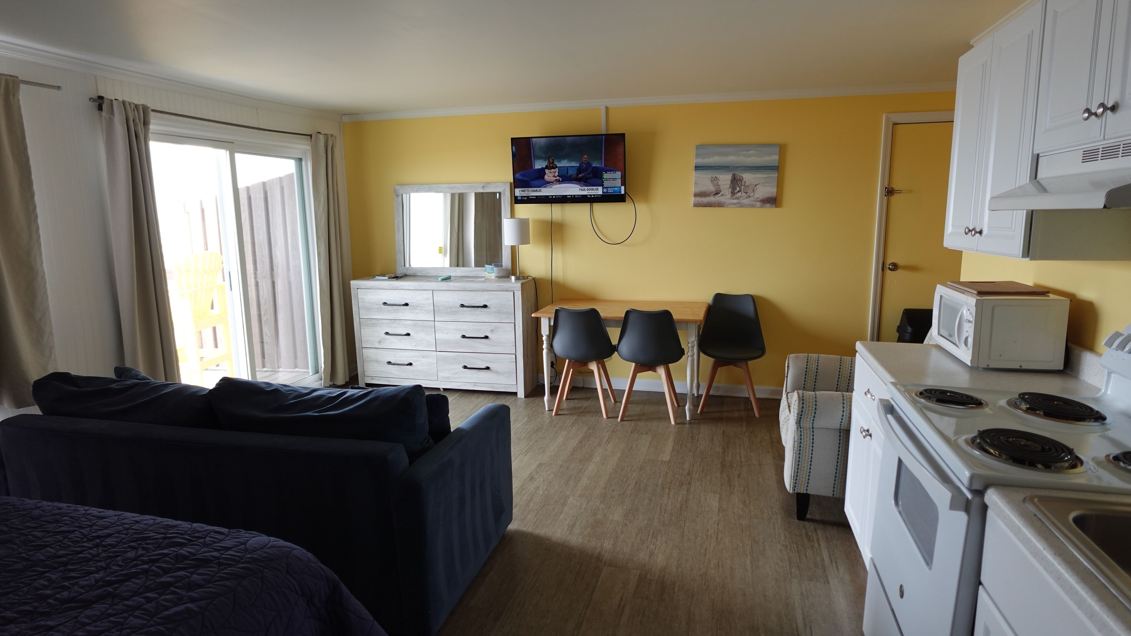 A compact hotel room with a bed, a small dining area with a table and chairs, a microwave, a stove, a television, and a window with curtains.