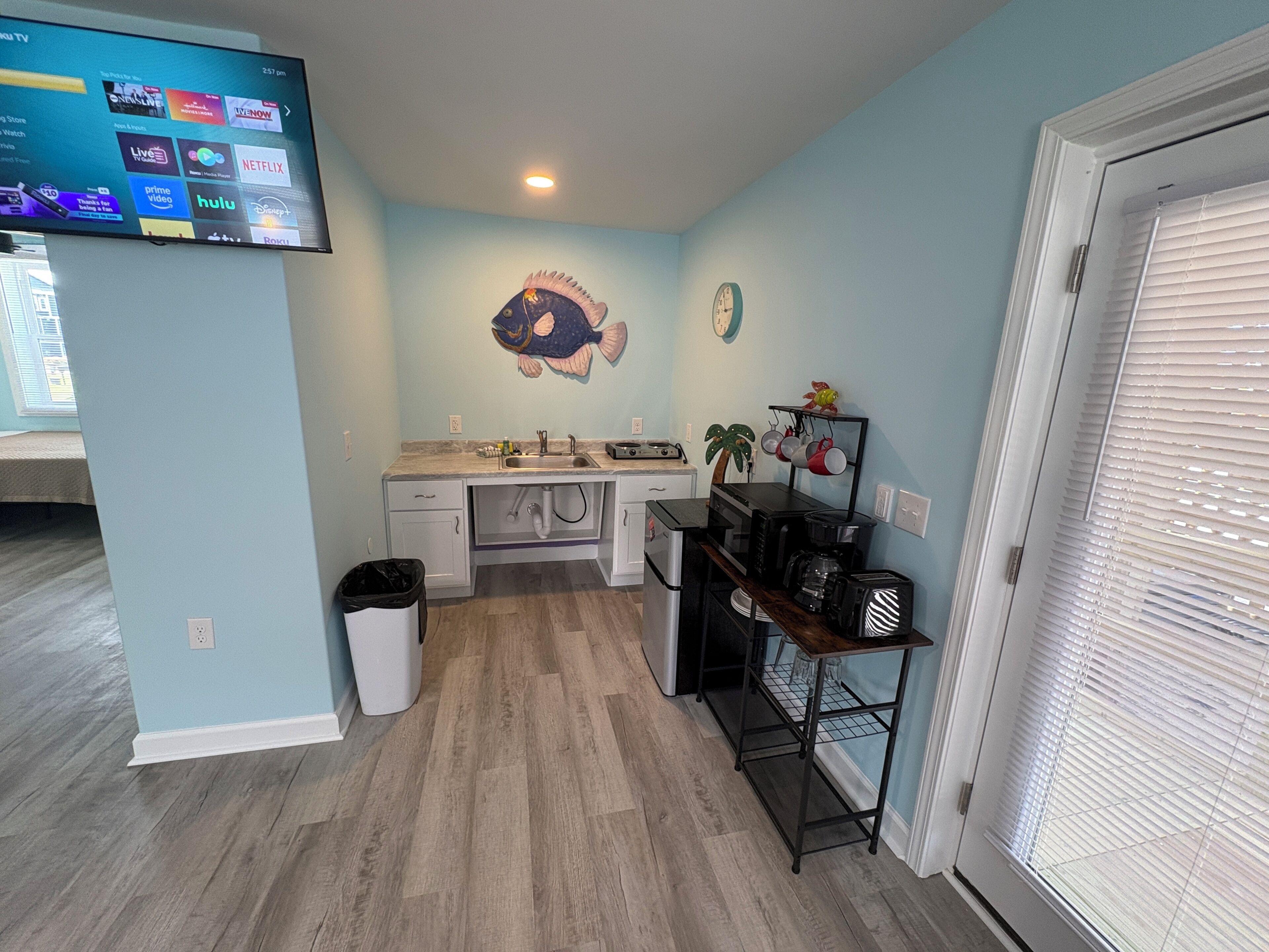 A compact kitchen with a sink, microwave, and coffee maker, a fish mural on the wall, and a TV mounted above the counter.