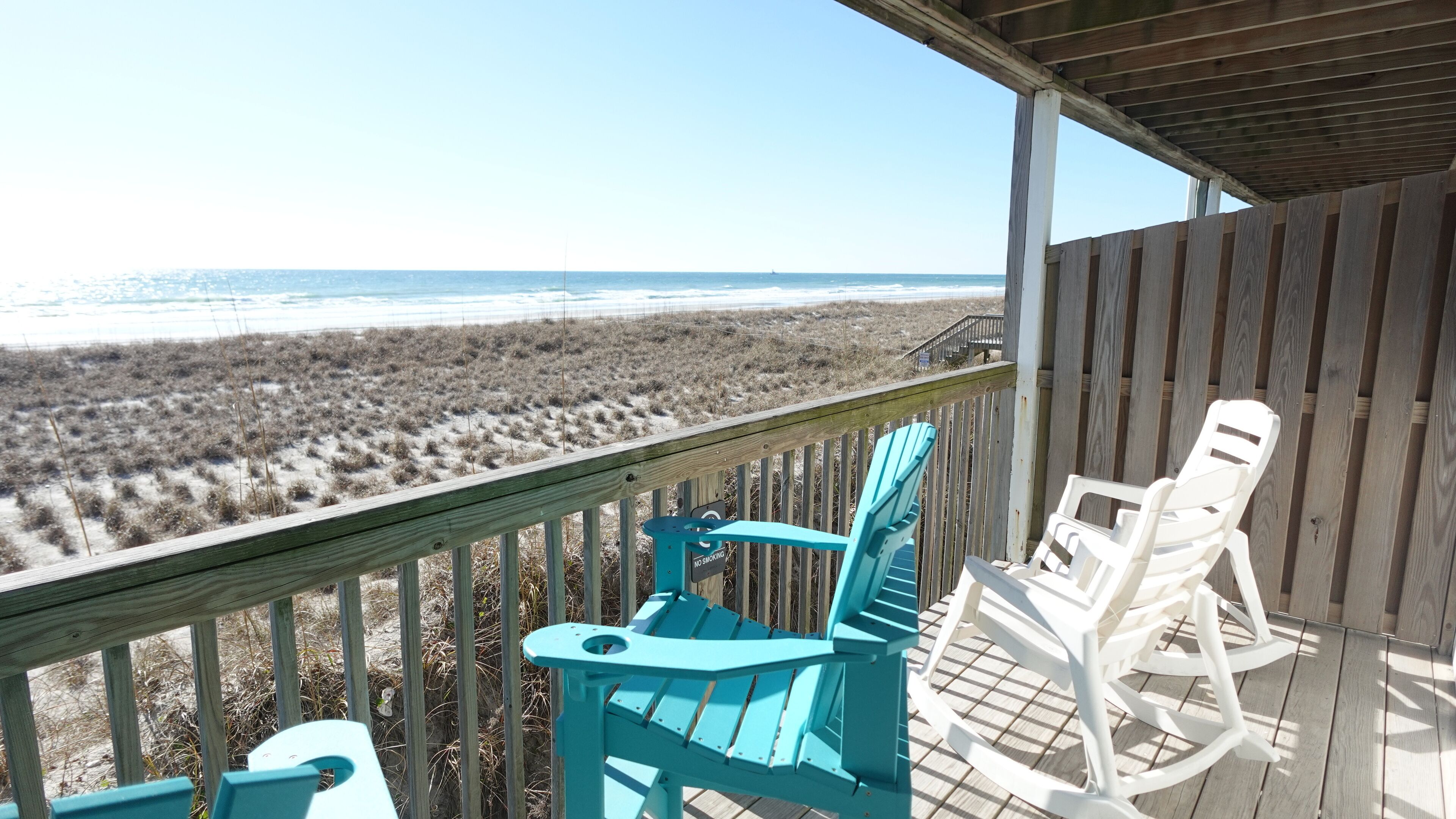 Une terrasse en bois avec deux fauteuils à bascule et une table, donnant sur une plage bordée de dunes et sur l’océan.
