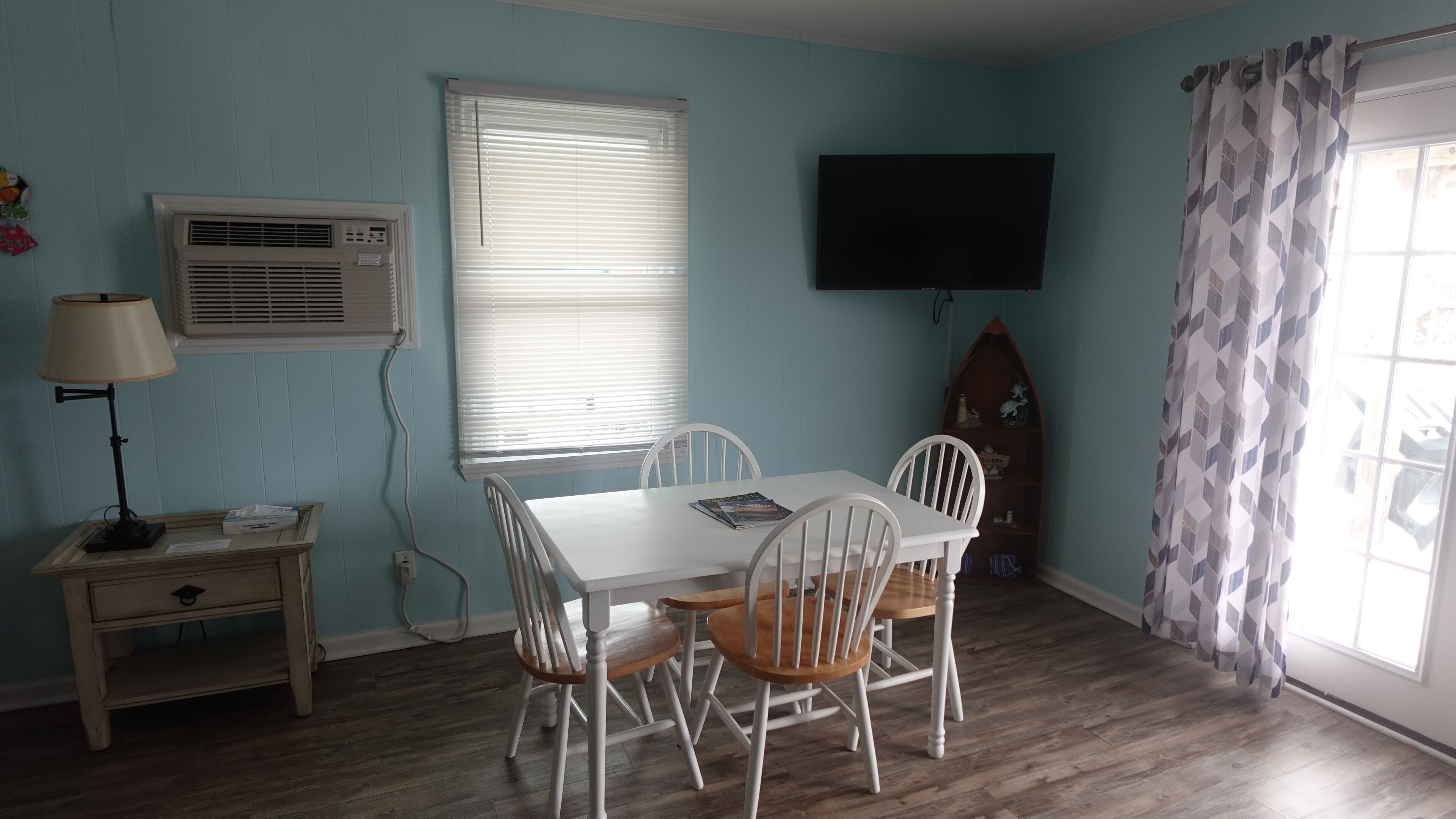 A room with a white table and chairs, a TV mounted on the wall, a window with blinds, and a wooden floor.